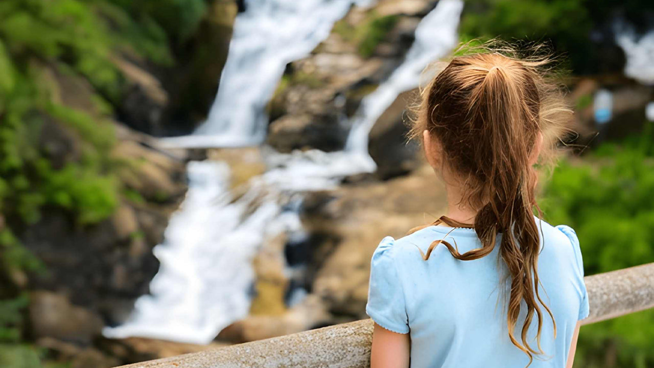 Girl in a light blue shirt sitting on a log looking at a Ravana waterfall in nature.